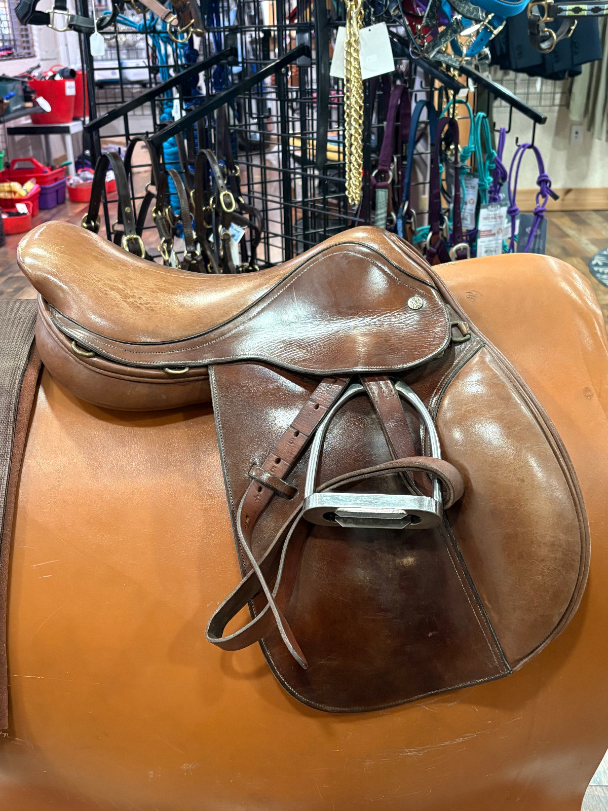 Brown leather saddle on display in a store with various equestrian equipment in the background.