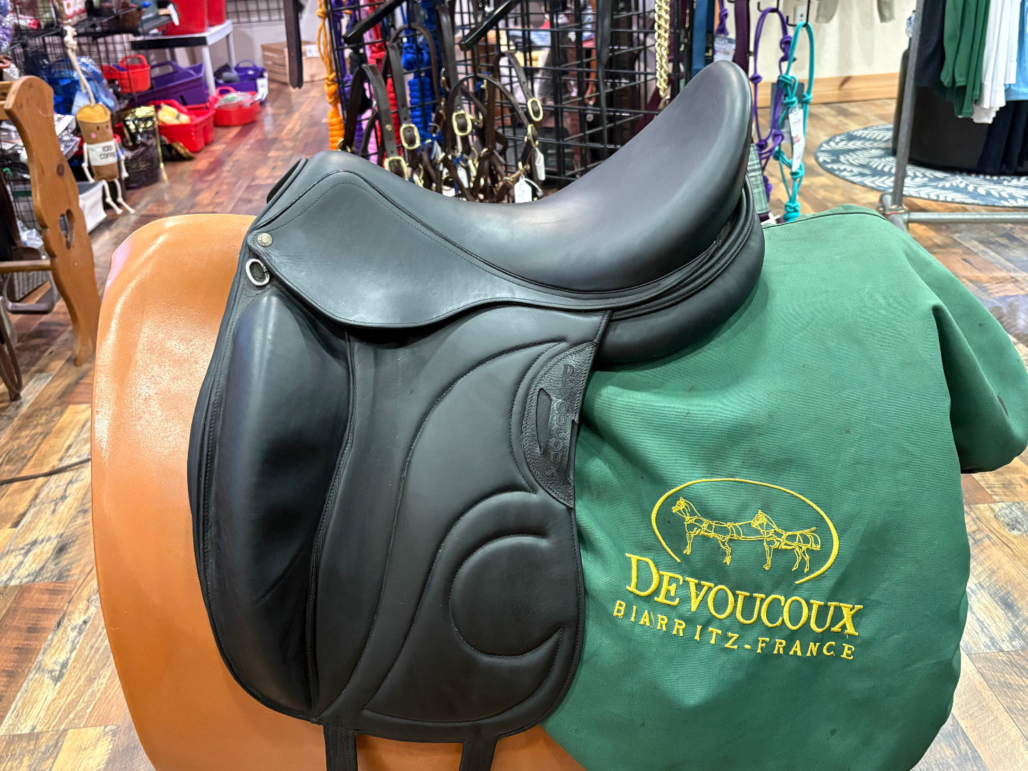 A black leather dressage saddle displayed on a stand, with a green Devoucoux Biarritz-France branded cover underneath the saddle, set in a retail store environment.