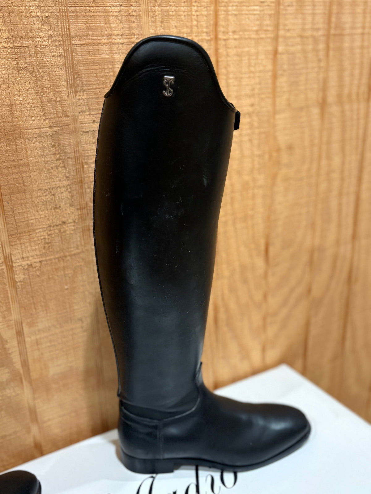 A single black leather equestrian riding boot standing upright on a white surface against a light wood-paneled background.