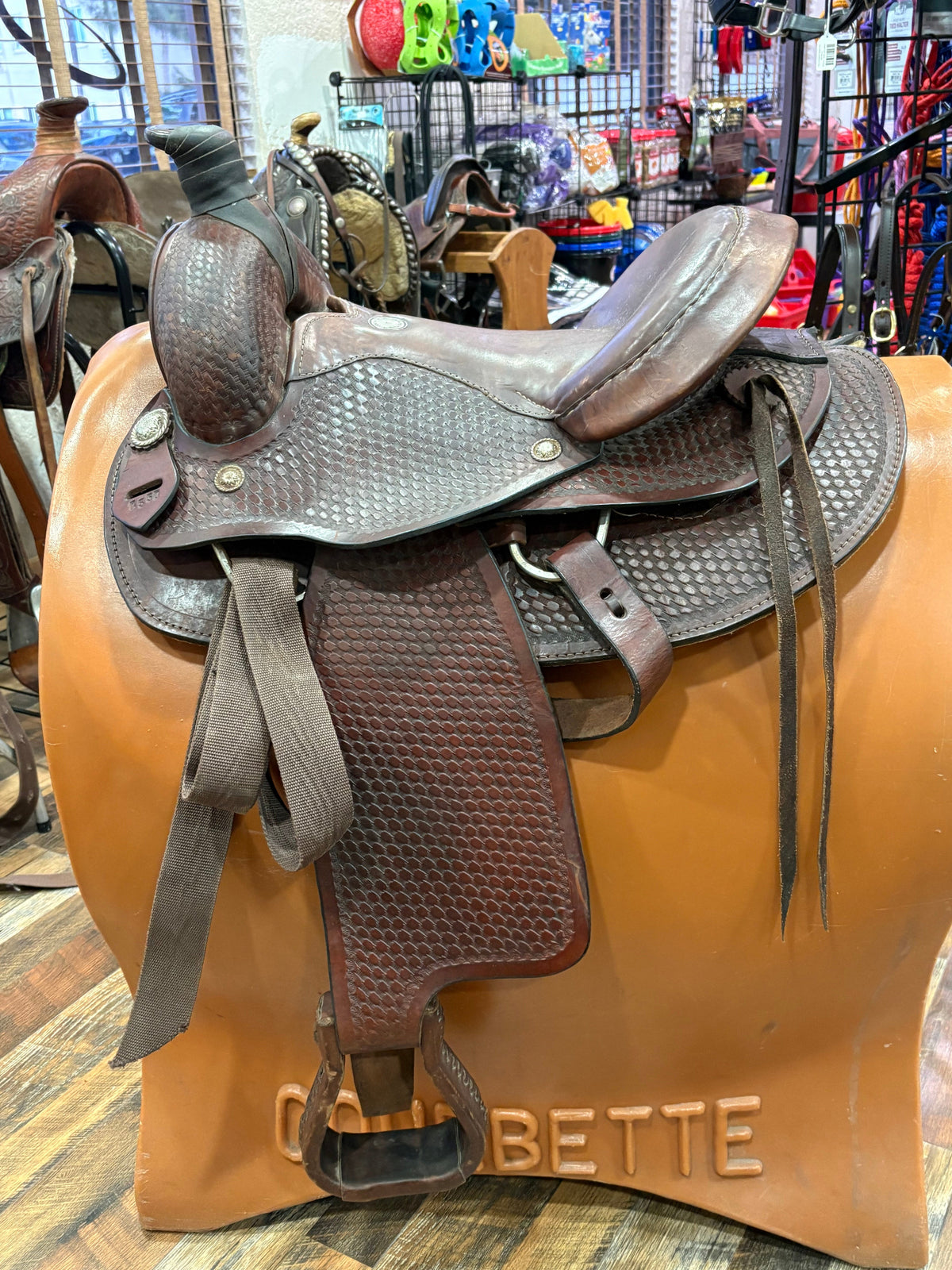 A brown leather western saddle with textured stitching, displayed on a tan stand inside a retail store.