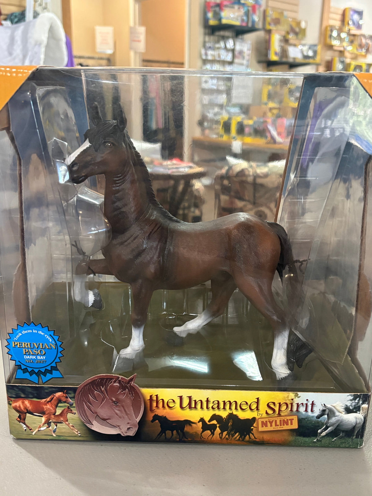 A brown horse figurine in a clear plastic display box with printed packaging, photographed in a retail store setting. The box shows the model name 'the Untamed Spirit' and 'NYLINT' branding.