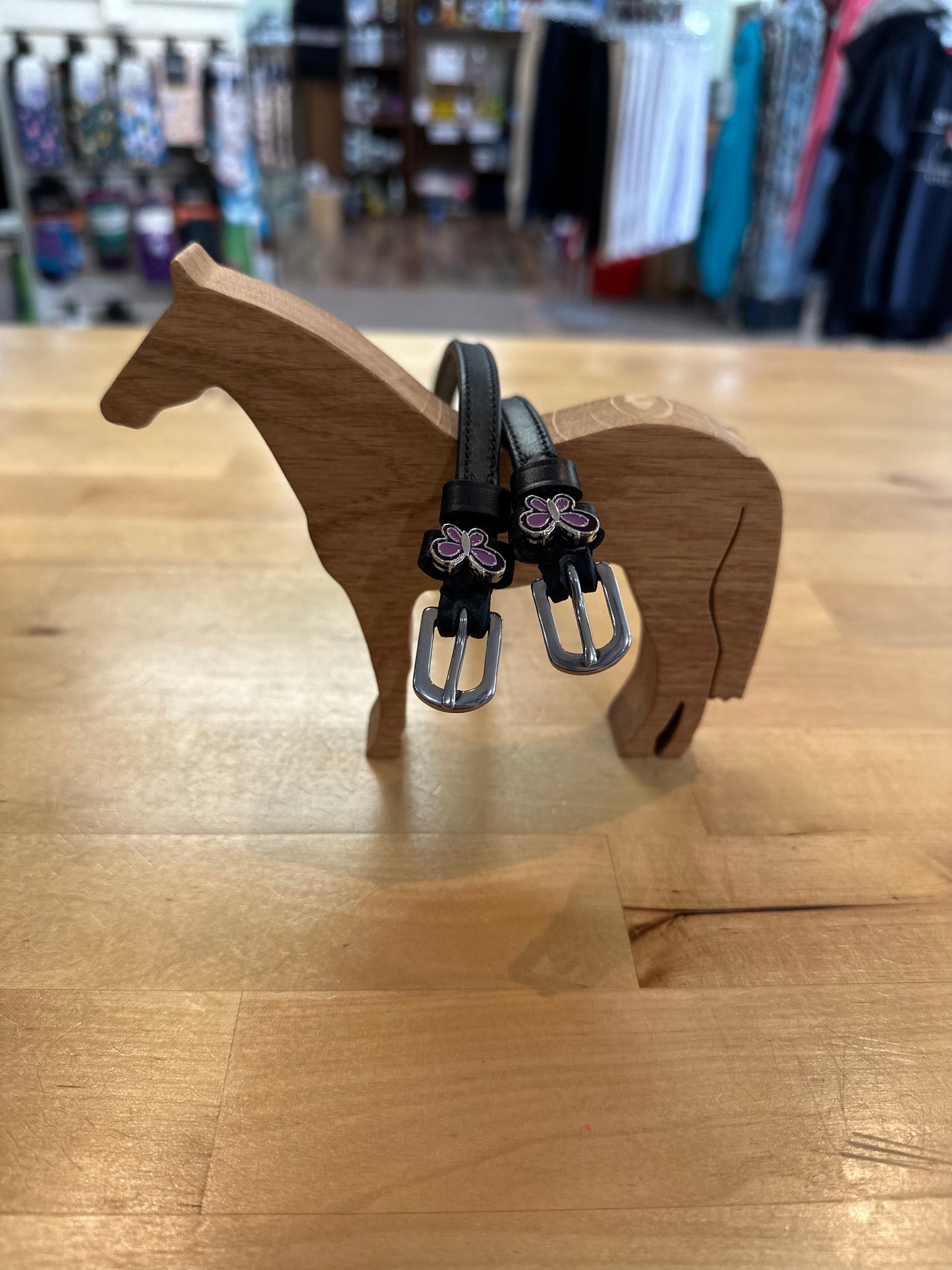 black leather equestrian spur straps with silver buckles with purple and silver butterfly charms resting on wooden horse model
