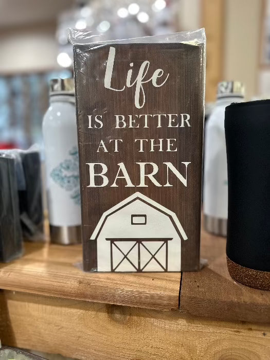 brown wood self sign with white words saying life is better at the barn, with a white decal of a barn