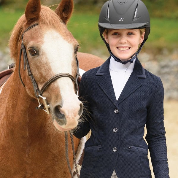 young girl dressed for a horse show in a navy show coat, white high collar shirt and black helmet. She is holding her bridled pony