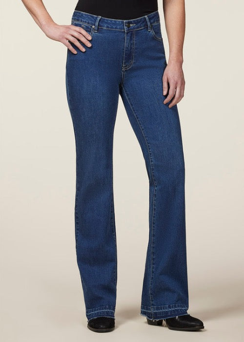 front view of lady wearing high-rise medium wash bootcut jeans with raw hem and wearing brown boots, and black tucked in shirt with right hand on hip standing against white background