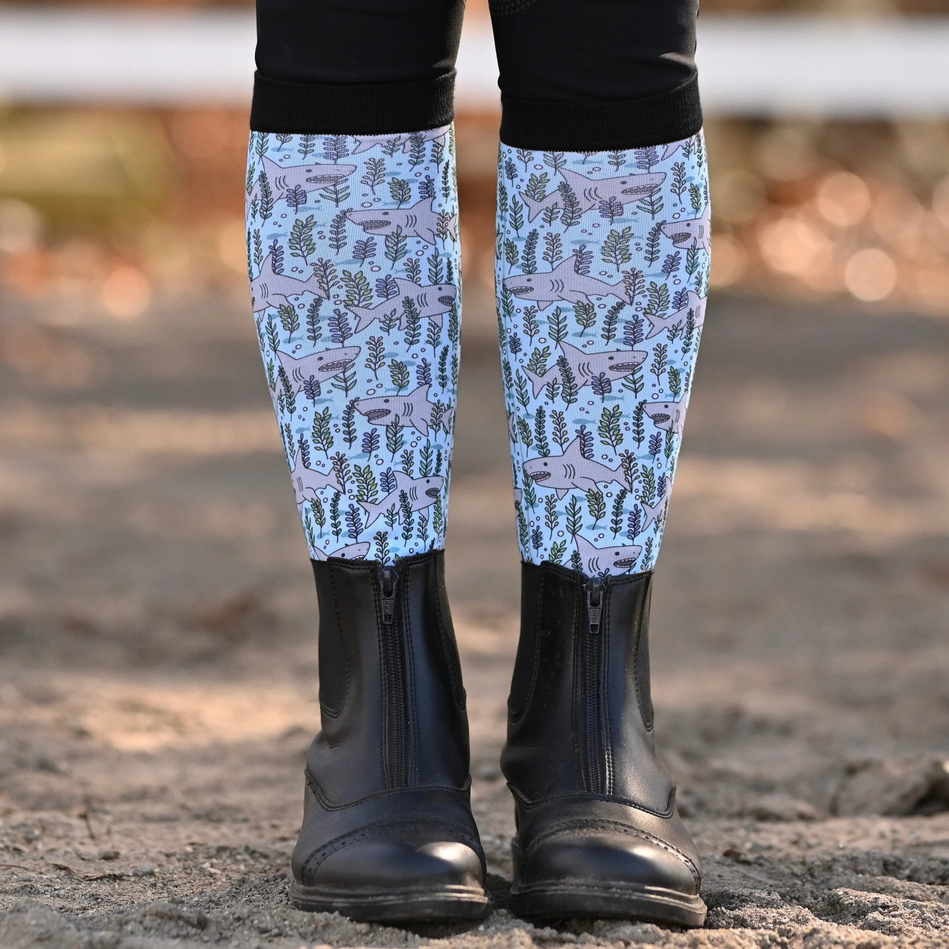Patterned boot socks with shark design worn by a person standing on a dirt surface.