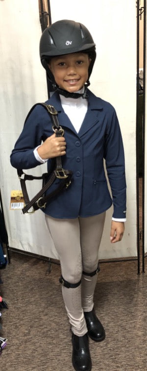 Little girl all dressed up and ready to go to a horse show. She's wearing tan jods with leather garter straps, black paddock boots, a navy show coat, white show shirt and an ovation helmet.
