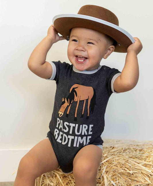 Child wearing a dark gray onesie with a horse graphic and text (Pasture Bedtime) sitting on straw.
