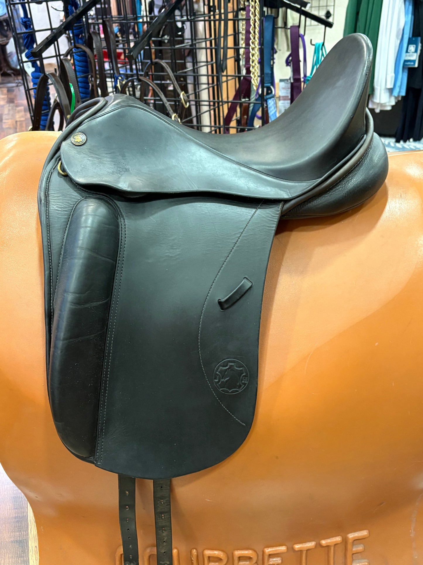 A black leather dressage saddle displayed on a stand, with a retail store interior visible in the background.