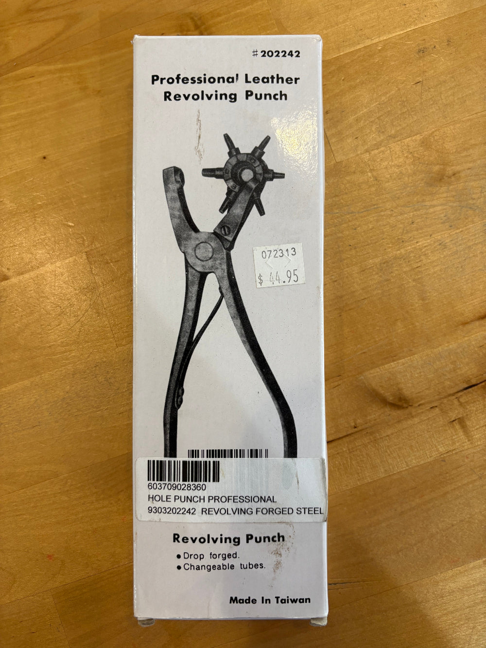 A boxed hole punch tool package on a wooden surface. The white box shows a black-and-white illustration of a leather revolving punch and text including 'Professional Leather Revolving Punch' 