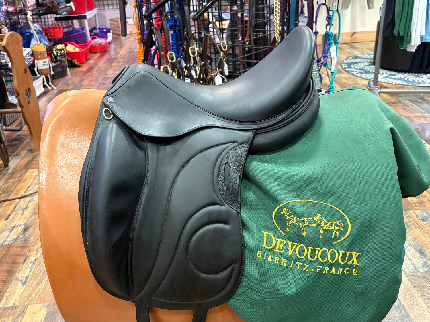 A black leather dressage saddle displayed on a stand, with a green Devoucoux Biarritz-France branded cover underneath the saddle, set in a retail store environment.