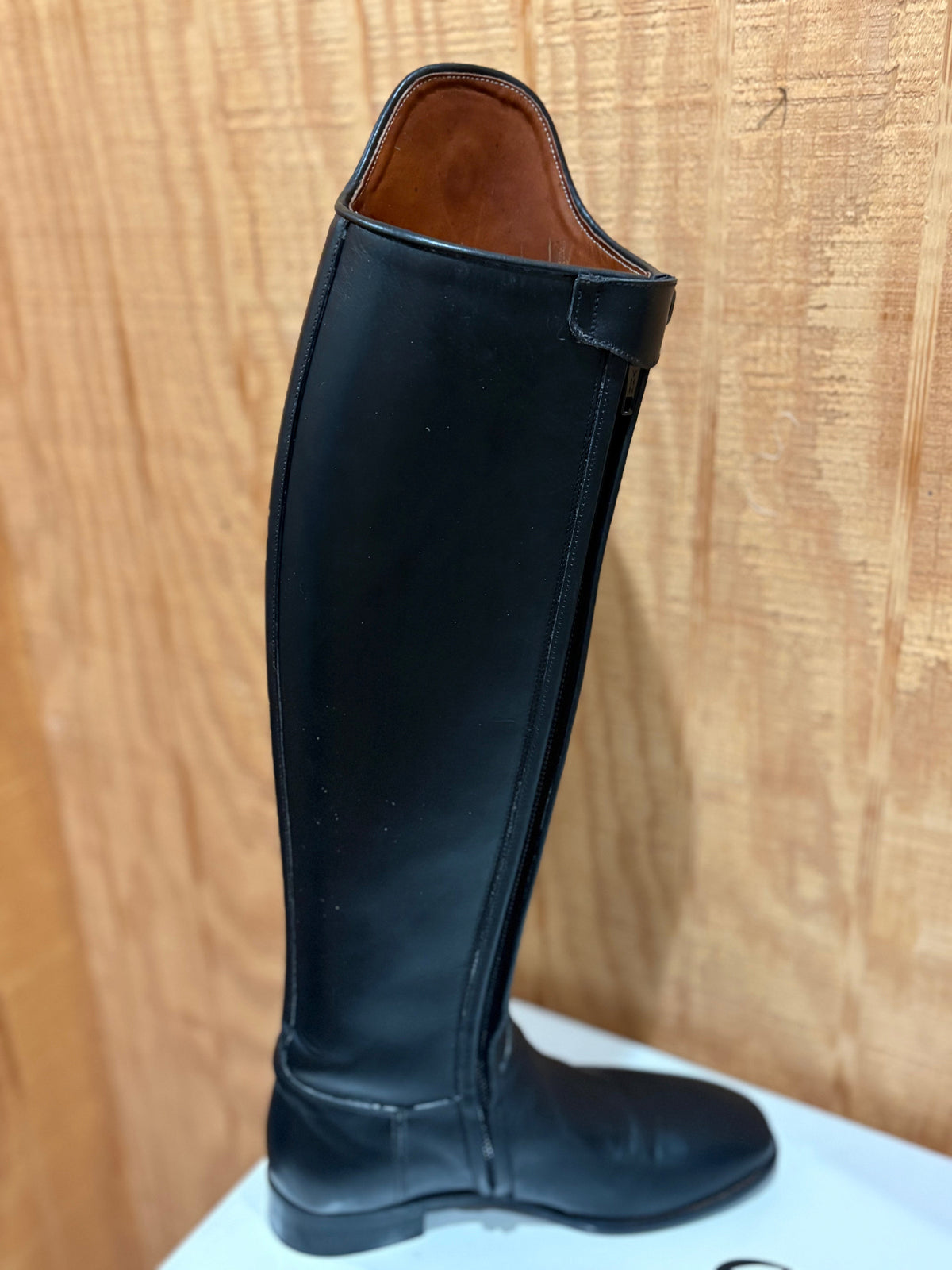 A single black leather knee-high riding boot shown in profile against a light wood panel background.