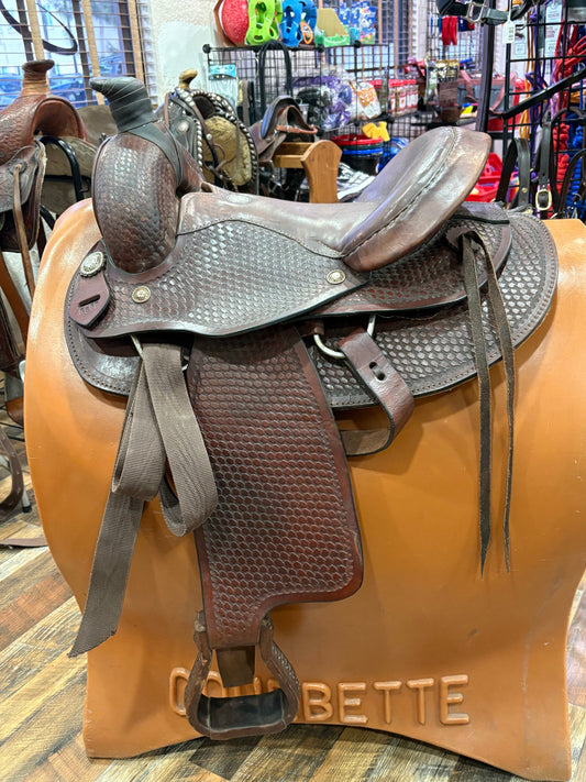 A brown leather western saddle with textured stitching, displayed on a tan  stand inside a retail store.
