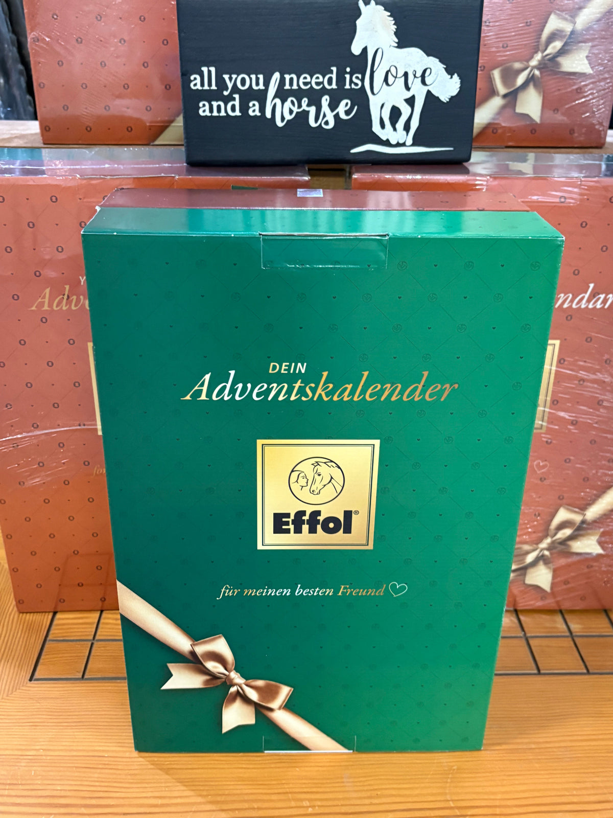 A green rectangular gift box for an Adventskalender by Effol, featuring gold lettering, a gold logo, and a brown ribbon bow on the front, photographed on a wooden surface with other gift boxes in the background.