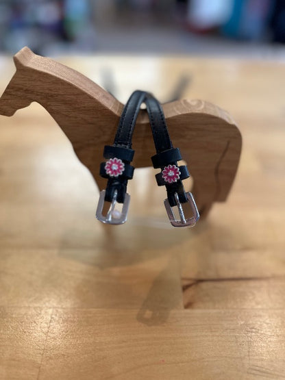 black leather equestrian spur straps with silver buckles with pink and silver flower charms resting on wooden horse model