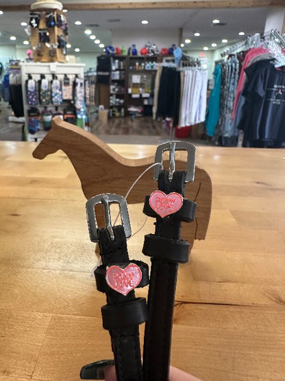 black leather equestrian spur straps with silver buckles with light pink and dark pink heart charm with the words pony love on it, resting on wooden horse model