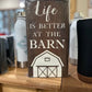 brown wood self sign with white words saying life is better at the barn, with a white decal of a barn