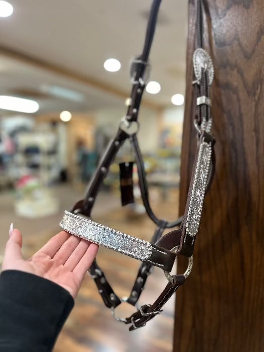 front view of brown and silver horse halter with hand holding base of halter with brown wood backdrop