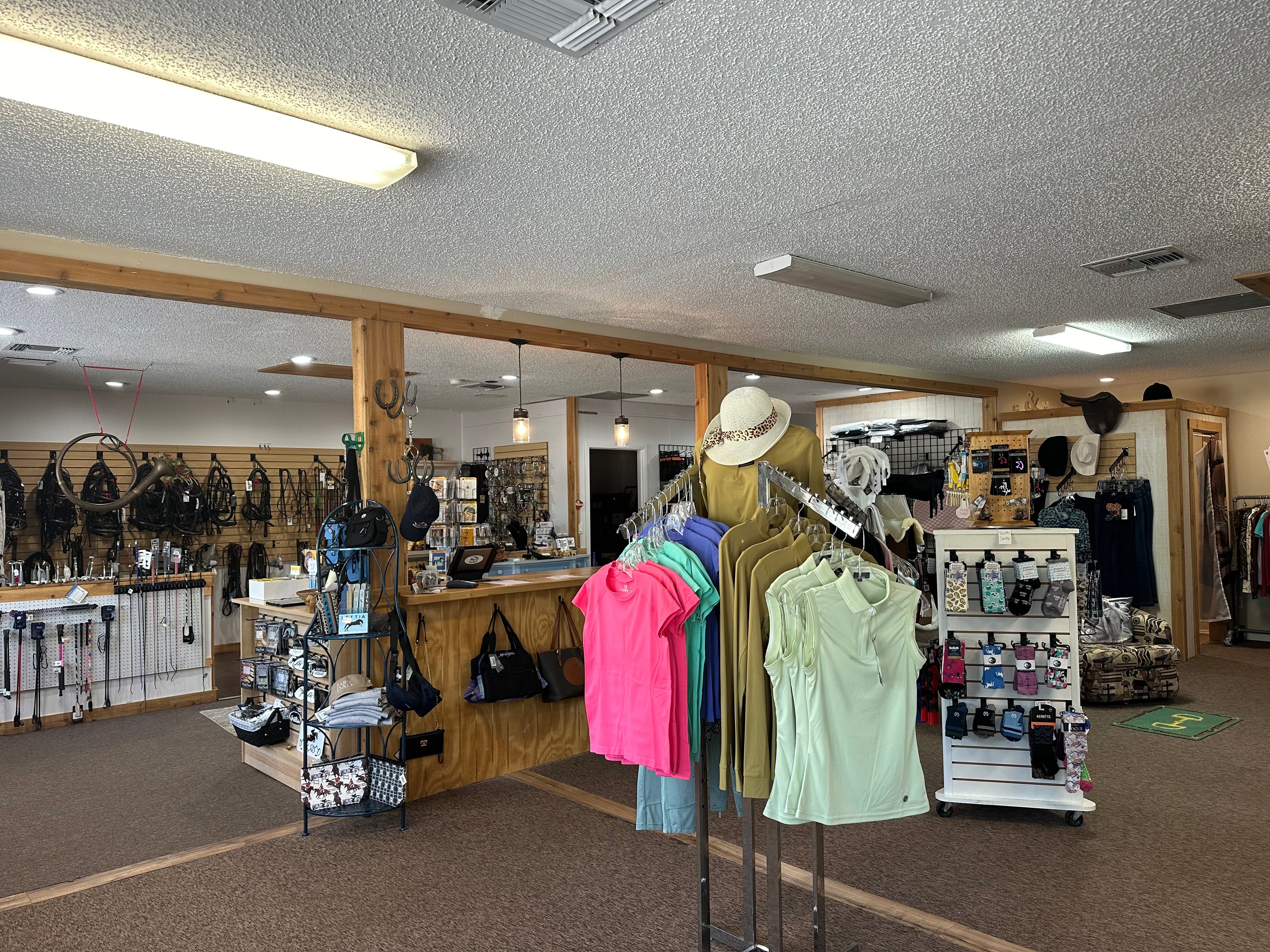 overview of shop to the front right rack of riding shirts behind that is the checkout counter, in the far left is a wall of bridles and to the far right there's saddle pads hanging up and socks on a rack next too the riding shirts