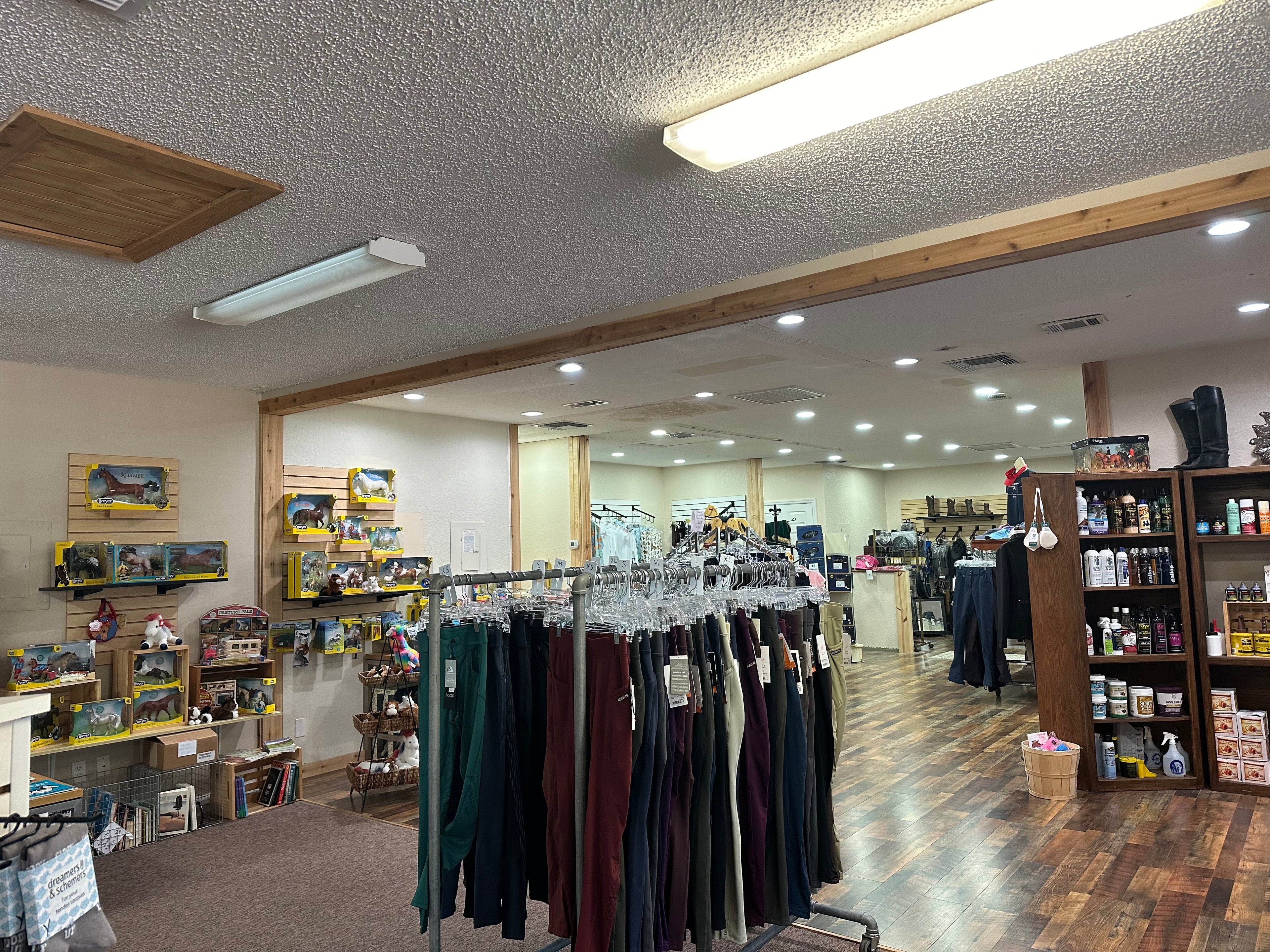 photo of overview of store pictured two racks of riding pants and on left wall boxes of horse figures and on the right shows horse soaps and in the far back youth clothing can be seen