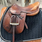 Light brown leather english style saddle with padded knee rolls and stirrups run up.  Saddle is on a stand with a black saddle pad.