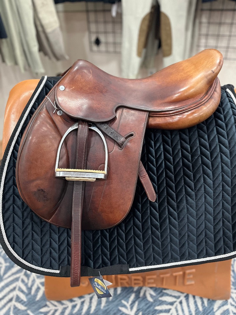 Light brown leather english style saddle with padded knee rolls and stirrups run up.  Saddle is on a stand with a black saddle pad.