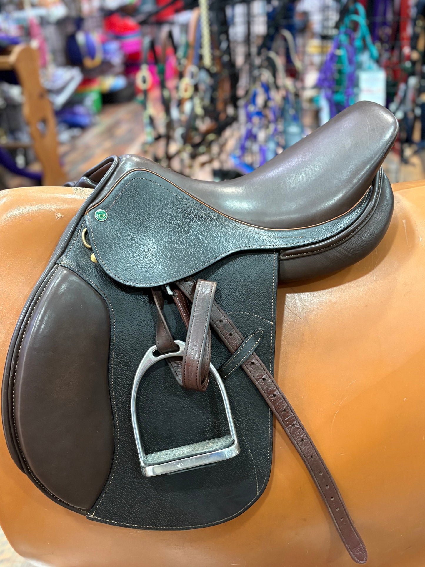 Brown leather english saddle on a saddle stand with a blurred background of colorful items.