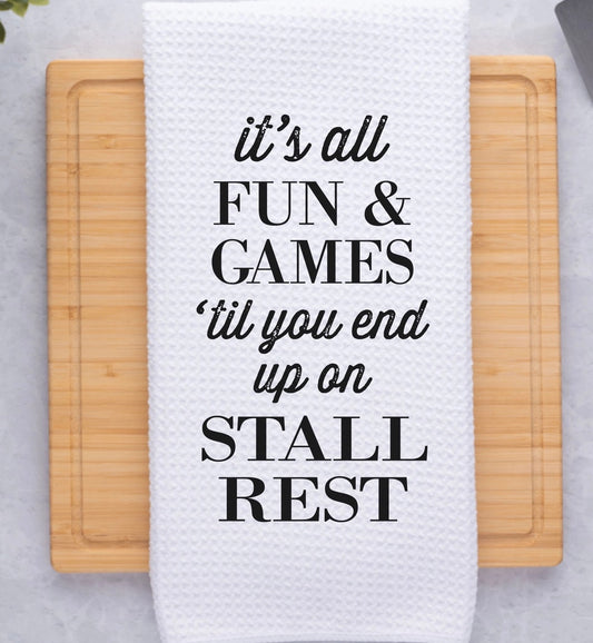 White waffle dish towel with the saying, "It's all fun & games 'till you end up on stall rest"