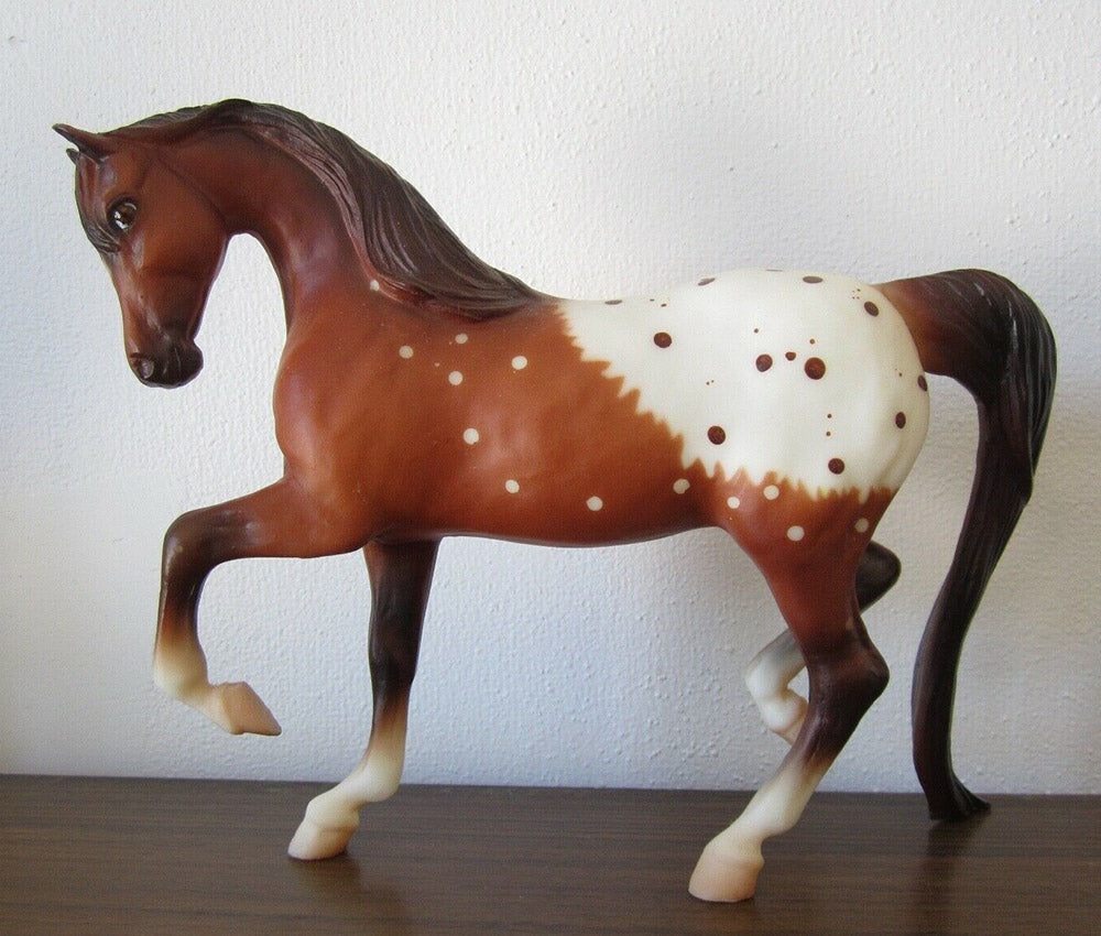 A painted toy horse figurine in a brown and white spotted pattern, standing on a dark wooden surface against a white wall.