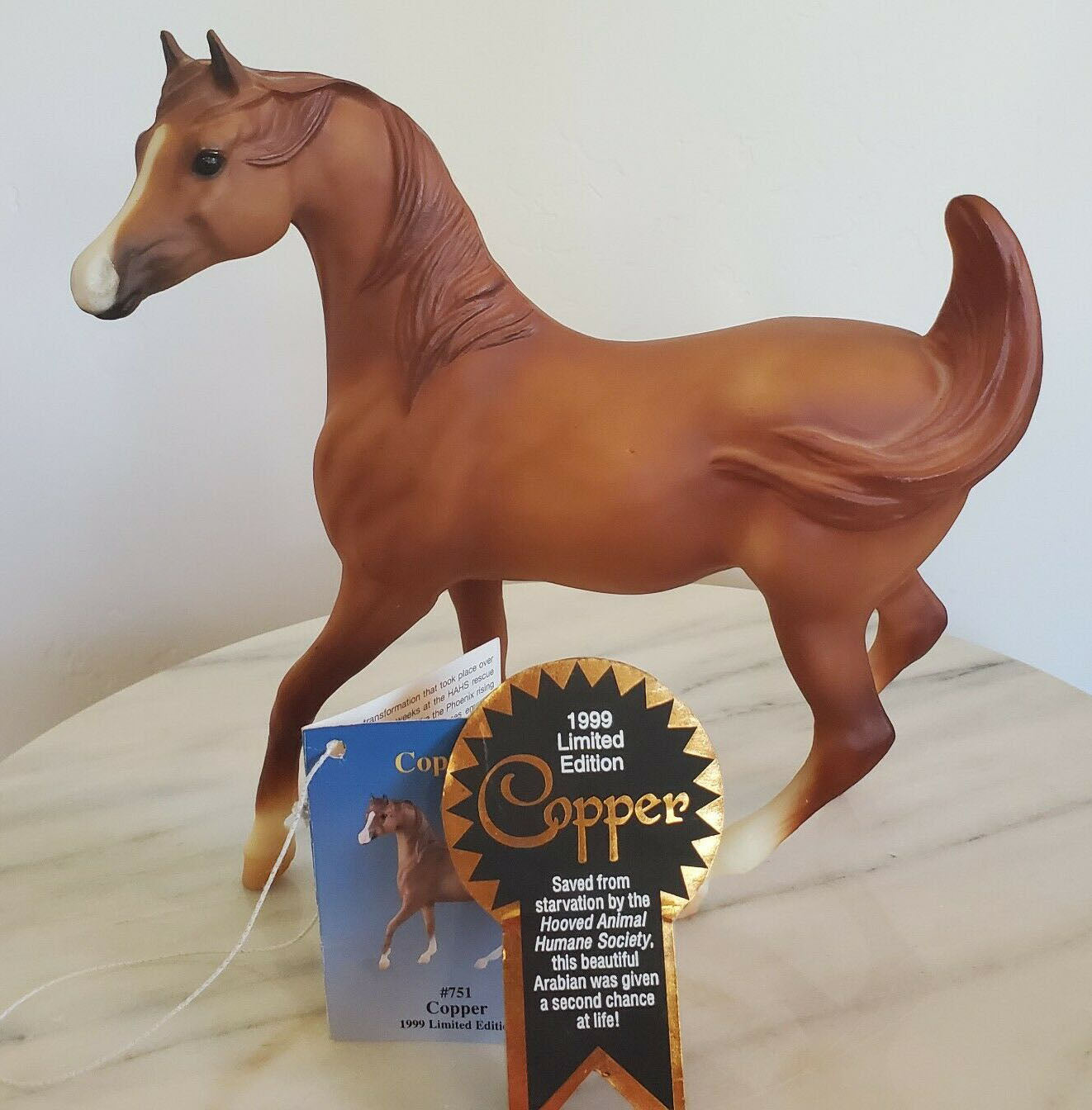A 1999 Limited Edition Breyer Copper horse figurine standing on a marble surface against a plain white wall, with its original blue card and a black and gold 1999 Limited Edition award ribbon in front.