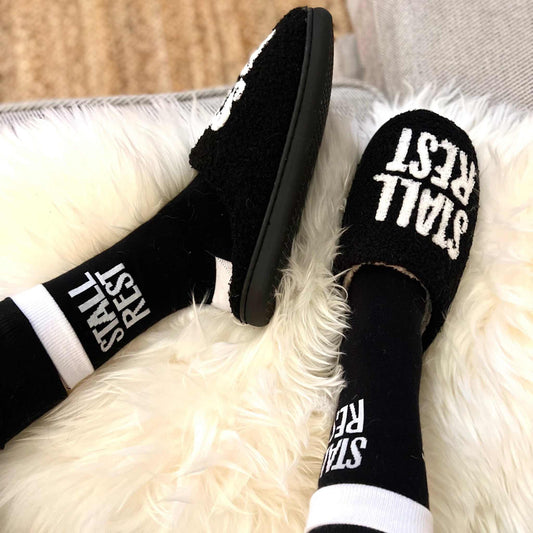 Black slippers with 'STALL REST' text on a fluffy white surface