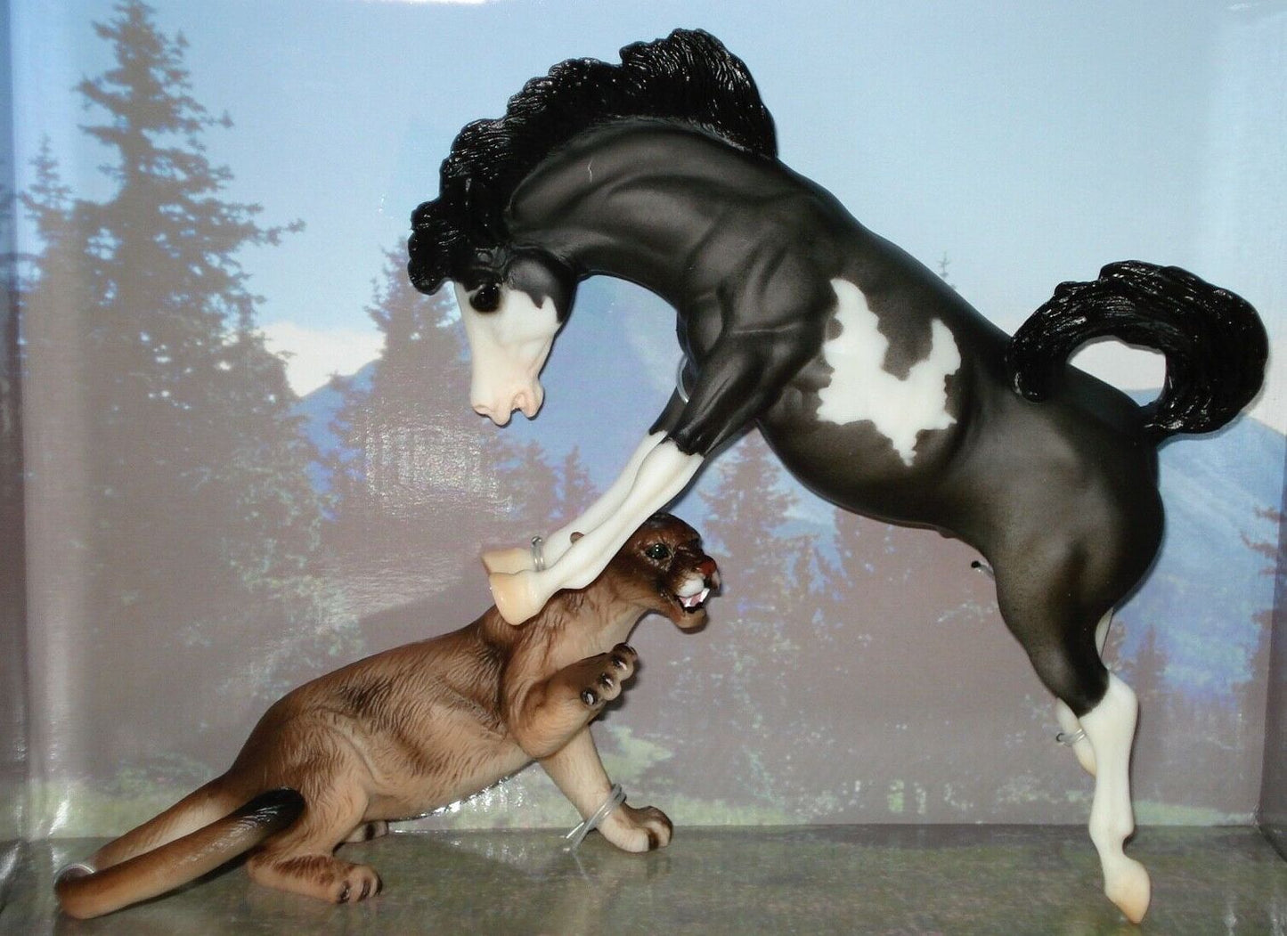 A toy figurine depicting a black and white horse rearing up to bite a brown mountain lion, set against a printed forest backdrop inside its original packaging.