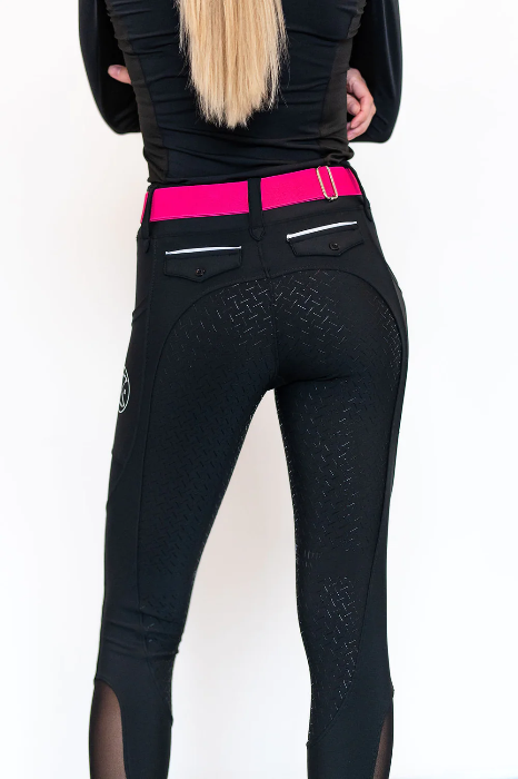 a blonde person standing away from the camera wearing black riding breeches with a hot pink belt 