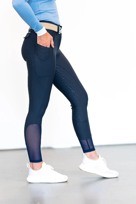 the right side of a person wearing navy riding breeches with hand halfway in side pocket with left food forward showing the inside of the pants with white tennis shoes