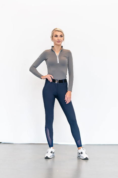 a blonde female facing forward with right hand on hip and left hand on left thigh wearing navy riding breeches with a long sleeve grey shirt and white and black tennis shoes 